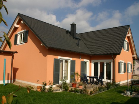 Ein pfirsichfarbenes Haus mit dunkelgrauem Dach und mehreren Fenstern mit Fensterläden verkörpert den Charme eines Dresdner Massivhauses. Der Terrassenbereich mit Tisch und Stühlen im Hinterhof ist von üppigem grünem Gras umgeben und liegt vor einem teilweise bewölkten Himmel.