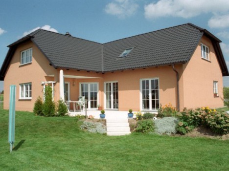Dieses zweistöckige Gebäude im Dresdner Massivhausstil mit pfirsichfarbenen Wänden und einem dunkelgrauen Ziegeldach ist von einem grünen Rasen und einem kleinen Garten umgeben. Es verfügt über mehrere Fenster und eine Terrasse mit Stufen, die zum Garten führen. Der Himmel ist klar mit ein paar Wolken.