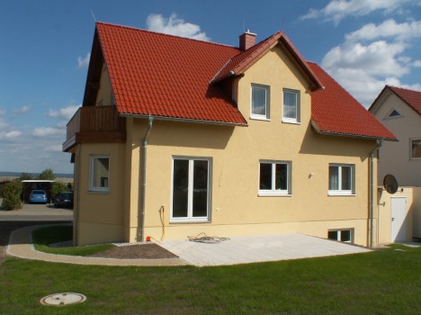 Ein zweistöckiges Haus mit gelber Außenfassade und rotem Satteldach, das an ein traditionelles Dresdner Massivhaus erinnert. Das Haus verfügt über mehrere Fenster und einen kleinen Balkon auf der linken Seite. Im Vordergrund erstrecken sich eine gepflasterte Terrasse und ein Rasen unter einem blauen Himmel mit Wolken.