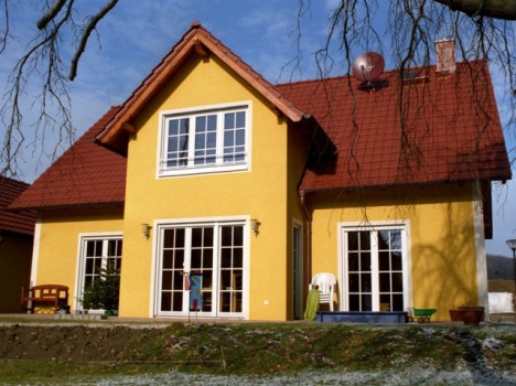Ein charmantes zweistöckiges gelbes Haus mit rotem Satteldach und großen Fenstern zeigt die Eleganz eines Dresdner Massivhauses. Ein kleiner Baum und Gartenstühle schmücken die Terrasse, während im schneebedeckten Hof, eingerahmt von nahe gelegenen Ästen, ein Spielzeuglastwagen steht.