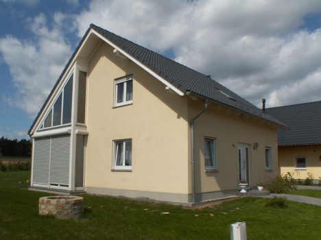 Ein beiges, zweistöckiges Dresdner Massivhaus mit einem dunkelgrauen Ziegeldach steht auf einem Rasengrundstück. Es verfügt auf einer Seite über große Glasfenster und eine weiße Eingangstür. Der Himmel ist teilweise bewölkt und ergänzt die charmante Architektur perfekt, ideal für alle, die einen Hausbau in Dresden in Erwägung ziehen.