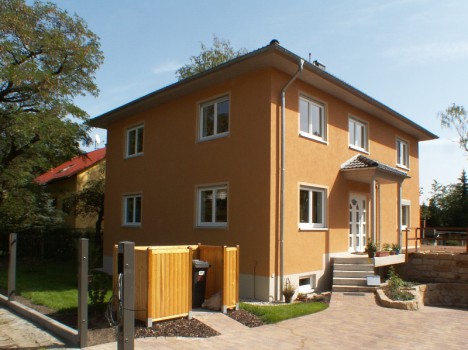 Ein zweistöckiges Haus, das die Eleganz eines Dresdner Massivhauses verkörpert, mit einer warmen beigefarbenen Fassade, weiß gerahmten Fenstern und einem Satteldach. Der Eingang verfügt über eine kleine Veranda mit Stufen, umgeben von einem makellosen Garten mit Bäumen und einer gepflasterten Auffahrt. Der Himmel ist klar und sonnig.