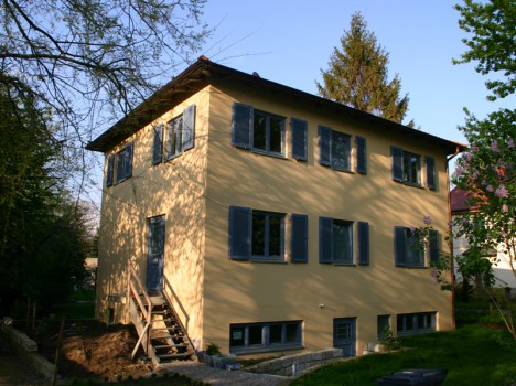 Ein zweistöckiges gelbes Haus mit blauen Fensterläden, typisch für die eleganten Massivhäuser Dresdens, ist von Bäumen umgeben. Eine kleine Treppe führt zum Vordereingang, wo das Sonnenlicht Schatten auf die Fassade des Hauses wirft.