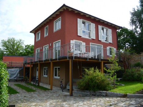Ein zweistöckiges rotes Haus mit weißen Fensterläden, typisch für Massivhäuser in Dresden, steht auf einem gepflasterten Platz, umgeben von Grün. Es verfügt über ein großes Oberdeck mit Geländer, Sitzgelegenheiten im Freien und mehreren Fenstern. Fahrräder können unten im Erdgeschoss abgestellt werden.