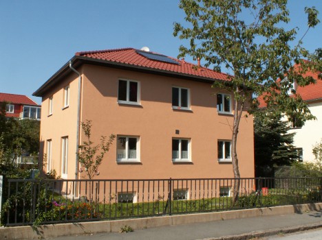Gezeigt wird ein zweistöckiges beiges Haus mit rotem Ziegeldach, das den eleganten Stil von Massivhäusern Dresden veranschaulicht. Davor steht ein Baum, der von einem schwarzen Metallzaun umgeben ist. Der klare blaue Himmel lässt auf einen sonnigen Tag schließen, perfekt, um die Handwerkskunst von Massivhausbau Dresden zu würdigen.