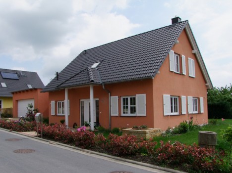 Ein pfirsichfarbenes Haus mit einem dunkelgrauen Ziegeldach und weißen Fensterläden steht an einer ruhigen Straße in Dresden. Vorne gibt es kleine Büsche und Blumenbeete. Der bewölkte Himmel unterstreicht die ruhige Vorstadtlage und spiegelt den Charme eines klassischen Dresdner Massivhauses wider.