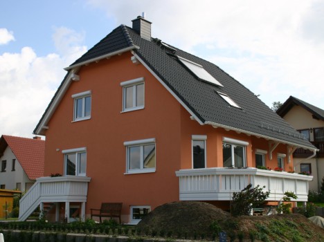 Ein charmantes zweistöckiges Haus im Stil eines Dresdner Fertighauses mit orangefarbener Fassade und dunkelgrauem Dach, akzentuiert durch weiß gerahmte Fenster und einen Balkon. Der Hof umfasst einen kleinen Garten und einen Erdhügel unter einem teilweise bewölkten Himmel, im Hintergrund sind die umliegenden Häuser zu sehen.