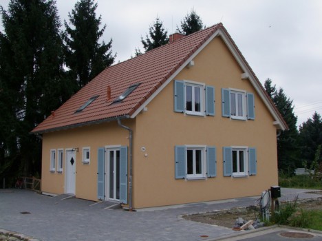Ein charmantes zweistöckiges Massivhaus in Sachsen mit hellbrauner Außenfassade und rotem Ziegeldach. Es verfügt über mehrere weiß gerahmte Fenster mit blauen Fensterläden. Umgeben von Bäumen und einer gepflasterten Auffahrt ist es der Inbegriff von hochwertigem Hausbau in Dresden.