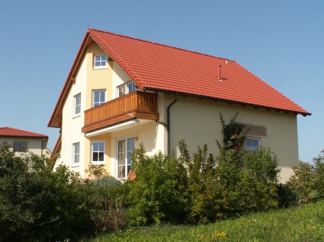 Dieses Dresdner Massivhaus ist ein charmantes zweistöckiges Haus mit einem roten Ziegeldach, einer hellgelben Außenfassade und einem Holzbalkon. Eingebettet in einen üppigen grünen Rasen mit Büschen und Blumen steht es stolz unter dem klaren blauen Himmel.
