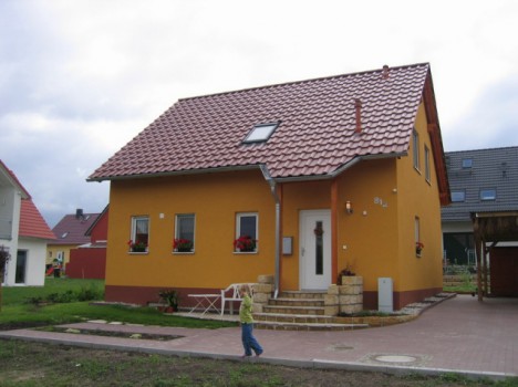 Ein kleines, orangefarbenes Haus mit rotem Ziegeldach und Topfblumen auf den Fensterbänken fängt den Charme der Massivhäuser Dresden ein. Ein Kind in einem grünen Hemd geht vorbei, und der Rasen und die gepflasterte Fläche verstärken den Reiz des Anwesens.