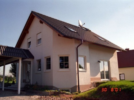 Ein charmantes zweistöckiges Haus mit hellen Wänden und einem dunklen Ziegeldach mit Oberlichtern und einer Satellitenschüssel. Dieses Dresdner Massivhaus verfügt über einen kleinen Eingang, der von einem Rasenhof und einem Kiesweg umgeben ist. In der unteren rechten Ecke ist das Datum „30 8 01“ zu sehen.