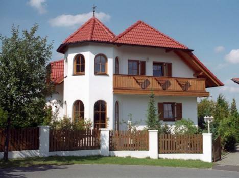 Ein zweistöckiges Haus mit weißen Wänden und einem roten Ziegeldach, typisch für ein Dresdner Massivhaus, mit Balkon und Bogenfenstern. Es ist von einem braunen Holzzaun und grünen Bäumen umgeben, vor einem blauen Himmel mit vereinzelten Wolken.