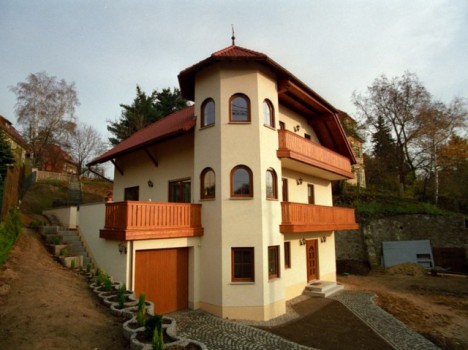 Dieses elegante dreistöckige Haus, das an ein Dresdner Massivhaus erinnert, verfügt über einen turmartigen Anbau und ein kastanienbraunes Dach. Es verfügt über gewölbte Fenster und Balkone mit Holzgeländern. Ein Steinpfad schlängelt sich durch eine üppige Landschaft, die sich wunderschön von einer von Bäumen gesäumten Umgebung und einer Steinmauer abhebt.