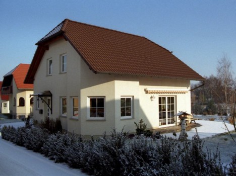 Ein gemütliches zweistöckiges Massivhaus in Sachsen mit rotem Ziegeldach, umgeben von einer verschneiten Landschaft. Das Haus verfügt über große Fenster und eine Holzterrasse. Im Vordergrund säumen schneebedeckte Sträucher den Gehweg. Ein klarer blauer Himmel vervollständigt diese Winterwunderlandszene.