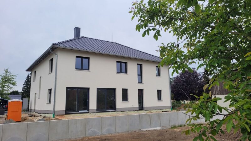 Ein modernes zweistöckiges Haus mit grauem Ziegeldach und cremefarbenen Wänden, umgeben von einem frisch angelegten Garten. Das Anwesen, das einem stilvollen Fertighaus in Dresden ähnelt, verfügt über große Fenster und einen sichtbaren Terrassenbereich. In der Nähe steht ein Baum mit grünen Blättern.