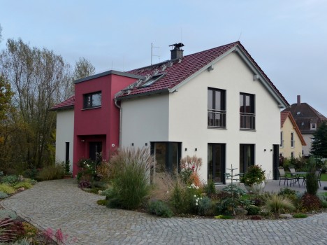 Ein modernes zweistöckiges Massivhaus in Sachsen verfügt über eine rot-weiße Fassade, große Fenster und ein mit roten Ziegeln gedecktes Schrägdach. Eine gepflasterte Auffahrt windet sich anmutig um einen angelegten Garten, der im Vordergrund mit Sträuchern und Ziergräsern geschmückt ist.
