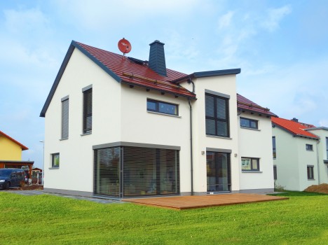 Modernes zweistöckiges Massivhaus Sachsen mit weißen Wänden, rotem Ziegeldach und großen Fenstern. Es verfügt über eine Holzterrasse im Hinterhof und eine Satellitenschüssel auf dem Dach. Der Rasen ist gepflegt und der Himmel ist klar und blau – ein idealer Rückzugsort im Dresdner Massivhaus.