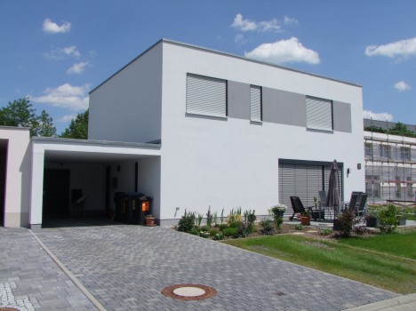 Modernes zweistöckiges weißes Haus mit Flachdach und großen Fenstern mit Fensterläden. Die Auffahrt führt zu einer offenen Garage. Sitzgelegenheiten im Freien mit Sonnenschirmen schmücken eine Terrasse, umgeben von einem gepflegten Garten und grünem Rasen unter blauem Himmel – der perfekte Rückzugsort im Massivhaus Sachsen.