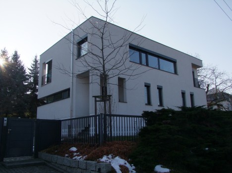 Ein modernes, minimalistisches zweistöckiges Haus mit Flachdach, großen Fenstern und heller Außenfassade ist ein Beispiel für die Eleganz von Massivhausbau Dresden. Umgeben von einem schwarzen Metallzaun und vereinzelten Bäumen, weicht ein Hauch von Schnee den Boden und den Bürgersteig auf.