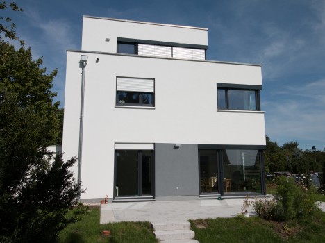 Ein modernes, weiß-graues, zweistöckiges Massivhaus in Sachsen mit großen Fenstern und einem Flachdach. Der Vorgarten verfügt über einen Steinweg, der von Büschen und Bäumen unter einem klaren blauen Himmel umgeben ist und die Eleganz des Hausbaus in Dresden zur Schau stellt.