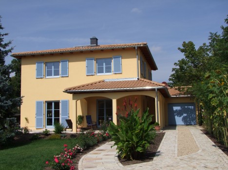 Ein zweistöckiges gelbes Dresdner Massivhaus mit blauen Fensterläden, einem Ziegeldach und einer Garage auf der rechten Seite. Es verfügt über eine Veranda, einen üppigen Garten mit roten und rosa Blumen und einen Steinweg, der zum Eingang führt.