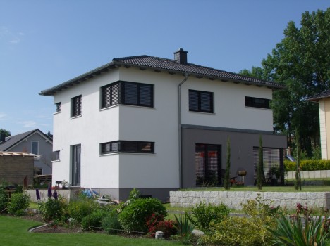 Ein modernes zweistöckiges weißes Haus mit grauem Dach und dunklen Fensterläden steht stolz da und zeigt den Charme des Massivhausbaus Dresden. Umgeben von einem gepflegten Garten mit üppigen grünen Pflanzen sonnt es sich unter einem klaren blauen Himmel, während im Hintergrund die Nachbarhäuser sichtbar sind.