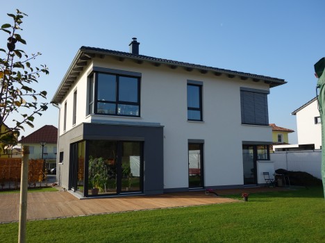 Modernes zweistöckiges Haus mit weißer und grauer Außenfassade im Stil der Dresdner Massivhäuser. Große Fenster auf beiden Etagen blicken auf einen gepflegten Rasen. Eine Holzterrasse auf der Rückseite ist mit Gartenmöbeln ausgestattet, alles vor der Kulisse eines klaren blauen Himmels.