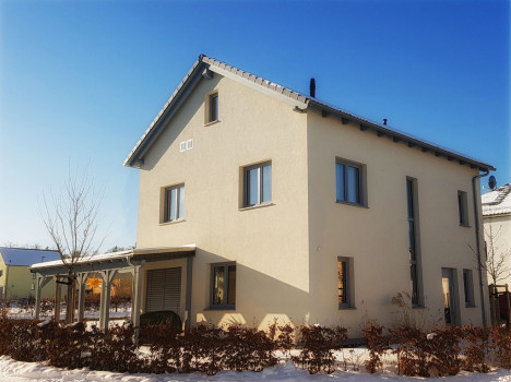 Ein zweistöckiges beiges Haus mit grauem Dach steht unter einem klaren blauen Himmel und erinnert an die charmanten Massivhäuser in Dresden. Schnee bedeckt den Boden und die umliegenden Büsche. Auf einer Seite befindet sich eine Veranda. Mehrere Fenster fangen das Sonnenlicht ein und verstärken den einladenden Reiz.
