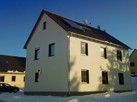 Gezeigt wird ein zweistöckiges Haus mit beigen Wänden und dunklem Dach, das den Charme der Massivhäuser Dresden verkörpert. Es verfügt über mehrere Fenster und Solarmodule auf dem Dach. Der Boden um dieses strukturell stabile Haus ist mit Schnee bedeckt. Links von ihm steht ein Auto unter einem klaren blauen Himmel.