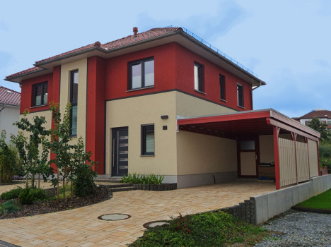 Dieses moderne zweistöckige Fertighaus in Dresden besticht durch eine rot-beige Außenfassade mit großen Fenstern und einem Flachdach. An der Seite ist ein hölzerner Carport angebracht, der den Charme noch verstärkt. Im Vordergrund ergänzen eine gepflasterte Auffahrt und ein angelegter Garten die Szene unter einem bewölkten Himmel.