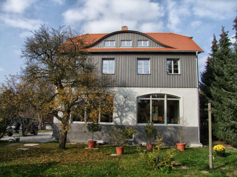 Ein zweistöckiges Massivhaus mit rotem Dach, grau-weißer Außenfassade und großen Fenstern steht elegant in der Landschaft. Umgeben von Bäumen, mit Topfpflanzen davor und einem Rasen. Der teilweise bewölkte Himmel verleiht diesem sächsischen Paradies seinen Charme.