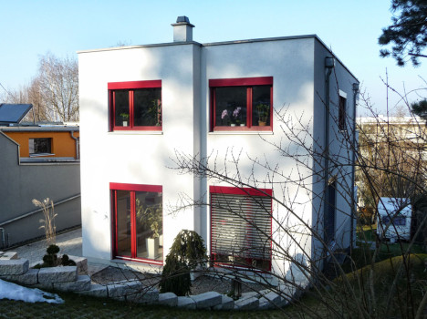 Ein modernes, zweistöckiges weißes Haus mit rotgerahmten Fenstern ist ein Zeugnis des Dresdner Massivhausbaus. Dieses elegante Gebäude ist von einem Garten mit kleinen Sträuchern umgeben, während ein Ast die Sicht teilweise versperrt. Der klare und sonnige Himmel vervollständigt diese malerische Szene.