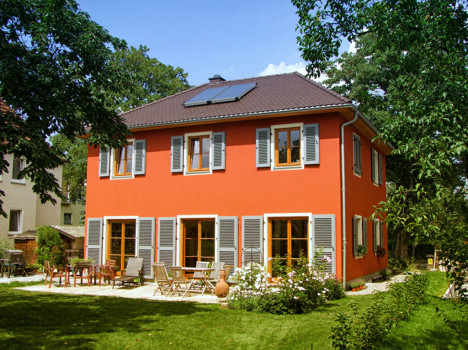 Dieses charmante zweistöckige, rote Dresdner Massivhaus verfügt über blaue Fensterläden und ein braunes Dach mit Sonnenkollektoren. Entspannen Sie auf der Terrasse mit Stühlen inmitten eines blühenden Gartens, umgeben von Bäumen, alles unter einem klaren blauen Himmel.
