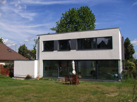 Dieses moderne, zweistöckige Dresdner Massivhaus besticht durch eine weiße Fassade und große Fenster. Es liegt auf einer weitläufigen Rasenfläche mit einem hölzernen Essbereich im Freien und zwei geschlossenen Sonnenschirmen. Hinter dem Haus steht ein hoher Baum unter einem teilweise bewölkten blauen Himmel und verkörpert die Essenz der Handwerkskunst der Dresdner Massivhäuser.