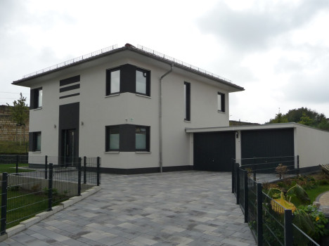 Ein modernes zweistöckiges Haus mit grau-weißer Fassade, großen Fenstern und einem Flachdach, typisch für den stilvollen Hausbau in Dresden. Es verfügt über eine Doppelgarage und eine geräumige gepflasterte Auffahrt, umgeben von einem niedrigen Zaun und einem gepflegten Garten. Der Himmel ist bedeckt.