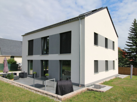 Modernes zweistöckiges Haus mit weißer Außenfassade und großen dunkelgrauen Fensterläden von Dresden Massivhaus. Es verfügt über einen Terrassenbereich mit Sitzgelegenheiten und Sonnenschirm, umgeben von einer Rasenfläche. Eine hohe Kiefer und benachbarte Häuser im Hintergrund veranschaulichen die Eleganz von Massivhaus Sachsen.