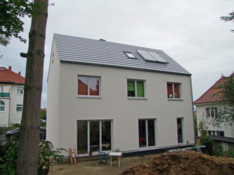 Ein modernes zweistöckiges Haus mit großen Fenstern und einem grauen Ziegeldach, auf dem ein Solarpanel angebracht ist. Die Außenfassade ist weiß, umgeben von einem Garten mit Tisch und Stühlen, der den Stil eines Dresdner Massivhauses inmitten teilweise sichtbarer Nachbargebäude widerspiegelt.