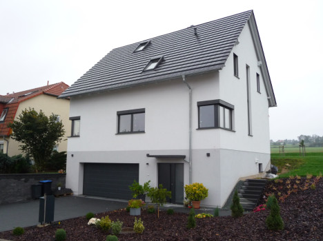 Ein modernes, zweistöckiges Dresdner Massivhaus mit grauem Ziegeldach und weißen Wänden. Es verfügt über große Fenster, eine Doppelgarage und einen angelegten Vorgarten mit Sträuchern und Blumen. Das Haus liegt auf einem Hanggrundstück, links ist ein Nachbarhaus zu sehen.