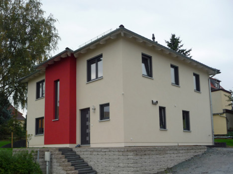 Dieses zweistöckige moderne Haus, ein schönes Beispiel für Massivhäuser in Dresden, verfügt über cremefarbene und rote Außenwände mit mehreren Fenstern und einer grauen Eingangstür. Es steht auf einem erhöhten Grundstück mit Steinstufen, die zum Eingang führen, und bietet eine malerische Aussicht, die durch die Bäume im Hintergrund noch verstärkt wird.