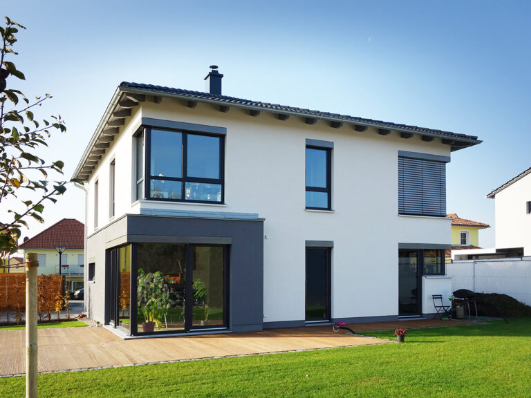 Modernes zweistöckiges Haus mit großen Fenstern, weißen Außenwänden, grauen Verkleidungen und einem Schrägdach. Das Haus ist von einem grasbewachsenen Rasen und einer Holzterrasse unter einem blauen Himmel umgeben.