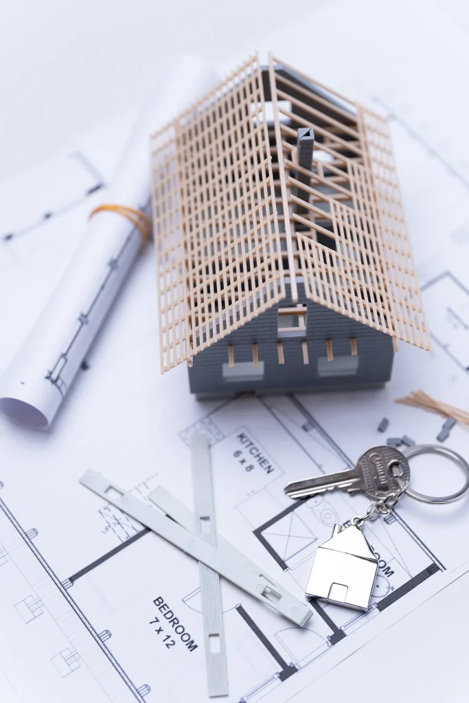 Ein maßstabsgetreues Modell eines Hauses mit einem hölzernen Dachstuhl liegt auf Architekturplänen, daneben ein aufgerollter Plan, ein Schlüsselbund mit einem hausförmigen Schlüsselanhänger und Messwerkzeuge.