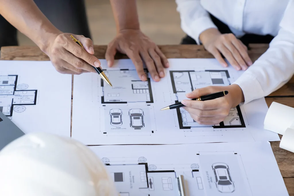 Zwei Personen begutachten und diskutieren die auf einem Holztisch ausgebreiteten Grundrisse von Häusern, wobei sie mit Stiften auf verschiedene Abschnitte der Baupläne zeigen.