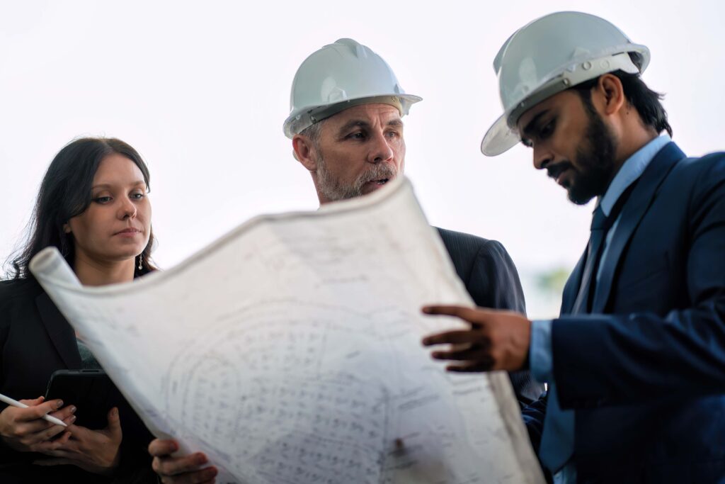 Drei Fachleute, zwei Männer mit weißen Schutzhelmen und eine Frau, betrachten gemeinsam eine große Blaupause. Der Mann auf der rechten Seite zeigt auf den Bauplan, während die anderen aufmerksam zuhören.