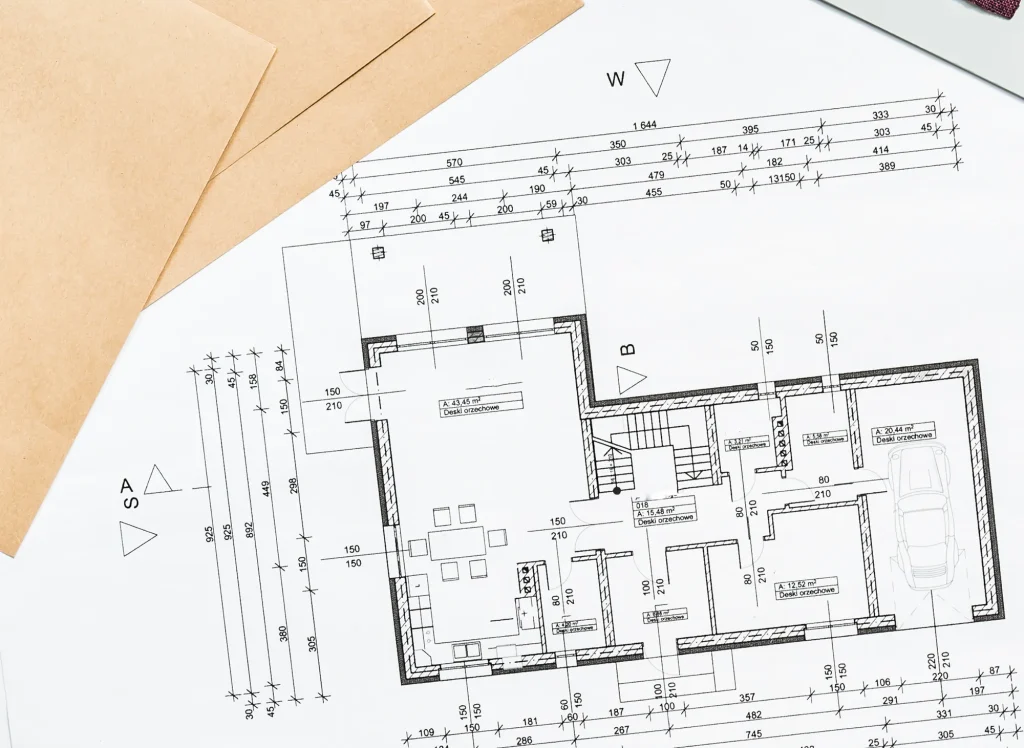 Ein detaillierter architektonischer Grundriss eines Hauses ist auf weißem Papier abgebildet, mit beschrifteten Räumen, Maßen und Symbolen. Braune Umschläge und ein Teil eines anderen Dokuments sind teilweise an der Seite sichtbar.