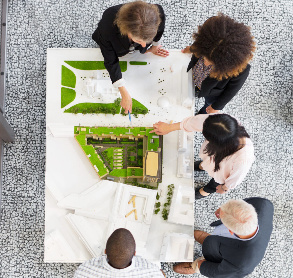 Fünf Personen stehen um einen Tisch herum und betrachten ein Architekturmodell mit Grünflächen. Sie diskutieren und zeigen auf verschiedene Bereiche des Entwurfs. Das Bild ist von oben aufgenommen.
