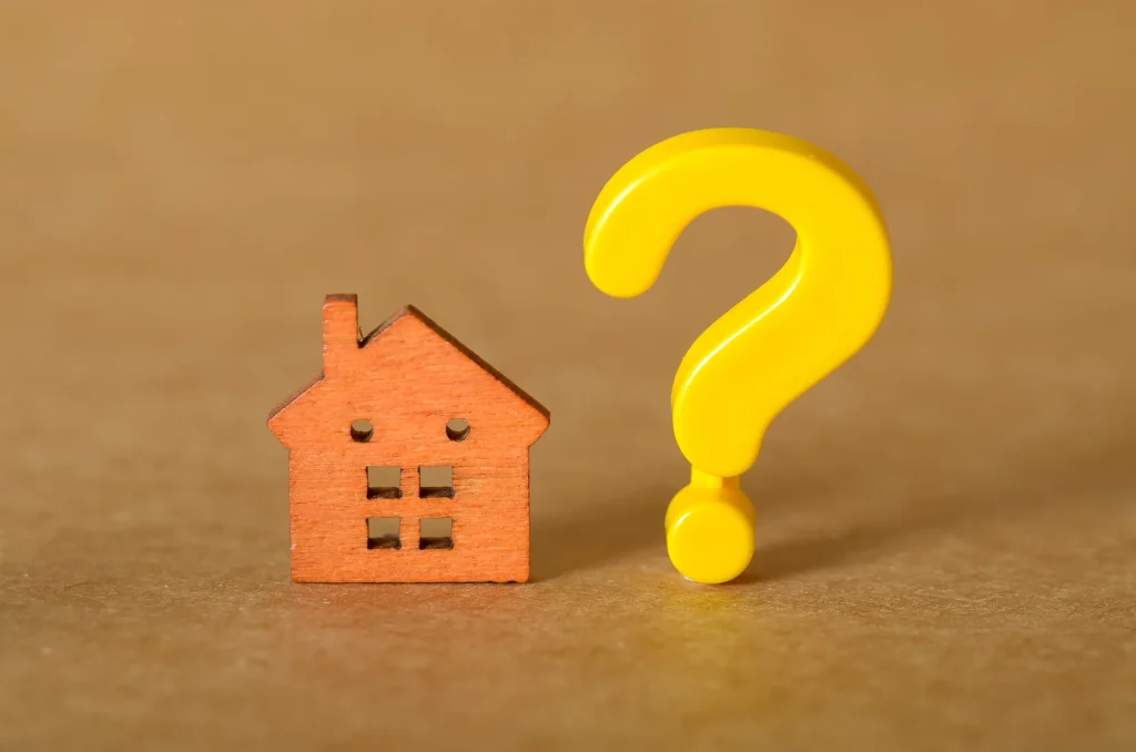 Eine kleine hölzerne Hausfigur steht neben einem großen gelben Fragezeichen auf einer braunen Fläche, das Fragen oder Unsicherheiten in Bezug auf Wohnraum oder Immobilien symbolisiert.