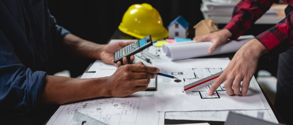 Zwei Personen prüfen Baupläne auf einem Schreibtisch mit einem Taschenrechner, einem Lineal und architektonischen Werkzeugen. Im Hintergrund sind ein gelber Schutzhelm und kleine Hausmodelle zu sehen.