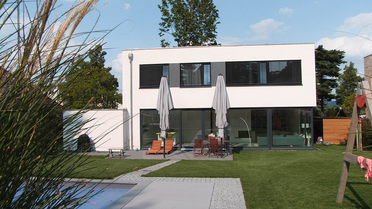 Ein modernes zweistöckiges Haus mit großen Fenstern, einem Flachdach und weißen Außenwänden, mit einer Terrasse mit Tischen, Stühlen und Sonnenschirmen, einem gepflegten Rasen und einem Swimmingpool im Vordergrund.