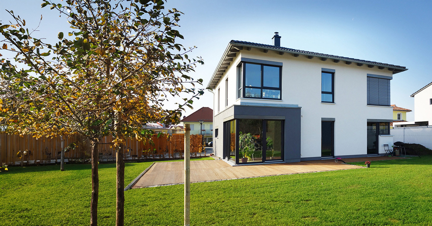 Ein modernes, zweistöckiges, weißes Haus im eleganten Stil von Hausbau Dresden steht auf einem weitläufigen, grünen Rasen mit großen Fenstern, einem jungen Baum im Vordergrund und einem eingezäunten Hinterhof unter einem strahlend blauen Himmel.