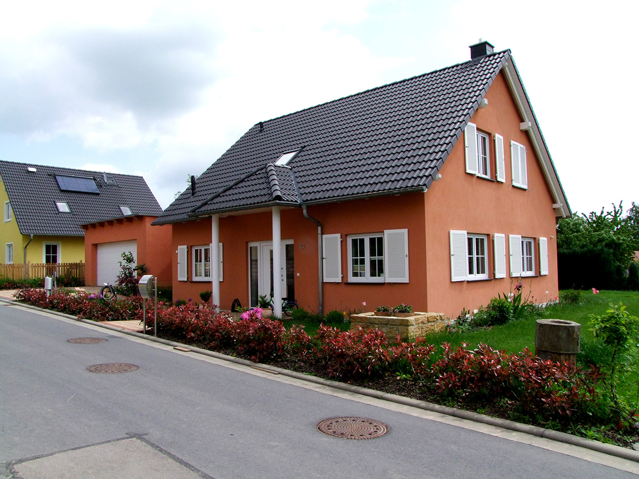 Ein pfirsichfarbenes Haus mit weißen Fensterläden und einem dunklen Ziegeldach steht in einer ruhigen Straße. Es gibt einen gepflegten Garten mit roten Sträuchern, grünem Gras und einer Einfahrt, die zu einer Garage in der Nähe führt.